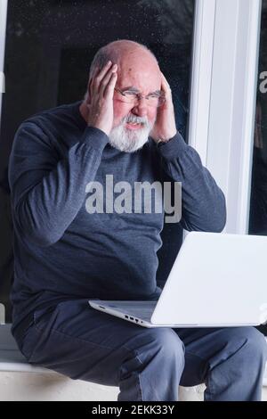 Ein älterer Mann, der nachts in seinem Wintergarten sitzt und versucht, einen Laptop zu benutzen. Stockfoto