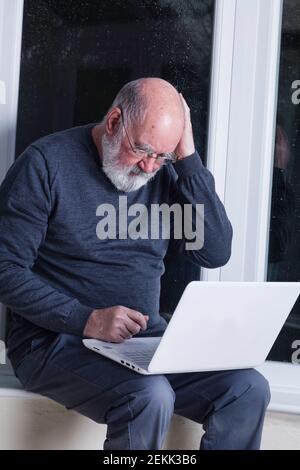 Ein älterer Mann, der nachts in seinem Wintergarten sitzt und versucht, einen Laptop zu benutzen. Stockfoto