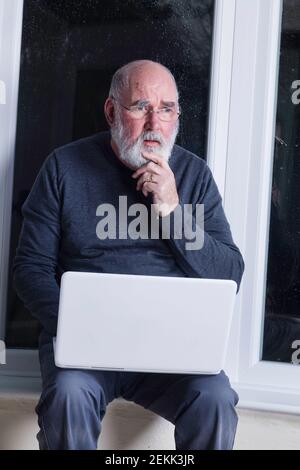 Ein älterer Mann, der nachts in seinem Wintergarten sitzt und versucht, einen Laptop zu benutzen. Stockfoto