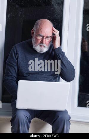 Ein älterer Mann, der nachts in seinem Wintergarten sitzt und versucht, einen Laptop zu benutzen. Stockfoto