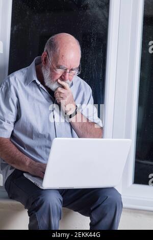 Ein älterer Mann, der nachts in seinem Wintergarten sitzt und versucht, einen Laptop zu benutzen. Stockfoto
