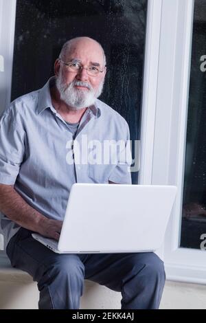 Ein älterer Mann, der nachts in seinem Wintergarten sitzt und versucht, einen Laptop zu benutzen. Stockfoto