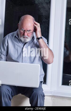 Ein älterer Mann, der nachts in seinem Wintergarten sitzt und versucht, einen Laptop zu benutzen. Stockfoto