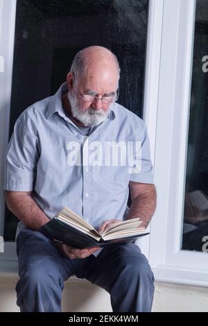 Älterer Mann, der nachts in seinem Wintergarten sitzt und einen Roman liest. Stockfoto