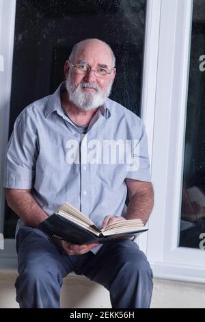 Älterer Mann, der in einer regnerischen Nacht in seinem Wintergarten sitzt und einen Roman liest. Stockfoto