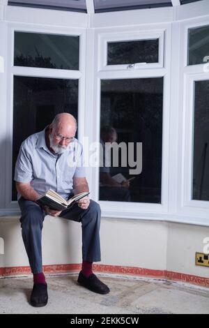 Älterer Mann, der in einer regnerischen Nacht in seinem Wintergarten sitzt und einen Roman liest. Stockfoto