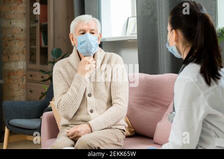 Kranker älterer Mann in Kasualwear und Maske sitzt auf der Couch, berührt seine Kehle und beschwert sich beim Arzt über Schmerzen und andere Symptome Stockfoto