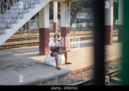 YANGON, MYANMAR - DEZEMBER 31 2019: Ein burmesischer Mann sitzt auf ein paar Reistüten und beobachtet sein Telefon an einem Bahnsteig des traditionellen Kreises Stockfoto