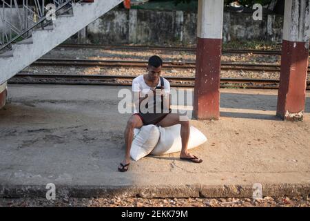 YANGON, MYANMAR - DEZEMBER 31 2019: Ein burmesischer Mann sitzt auf ein paar Reistüten und beobachtet sein Telefon an einem Bahnsteig des traditionellen Kreises Stockfoto