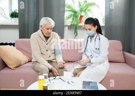 Junge Ärztin in Whitecoat und Schutzmaske mit Tonometer Während er neben kranken älteren Mann sitzt und sein Maß misst Blutdruck Stockfoto