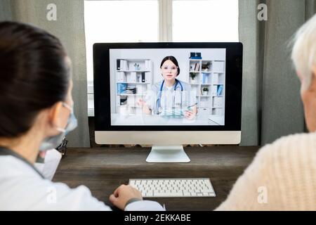Älterer Mann und Allgemeinpraktizierender, der vor dem Tisch sitzt Von Computer-Monitor mit jungen Ärztin auf dem Bildschirm während Beratung Stockfoto