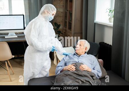 Junge Ärztin in Schutzhüllen berühren Schulter von Kranken Älterer Mann, der auf der Couch unter der Decke lag und ihm gab Medizinische Beratung Stockfoto