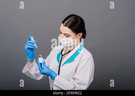 Seriöse Ärztin in einer medizinischen Maske hält Spritze und Impfstoff isoliert auf grau Stockfoto