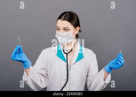 Seriöse Ärztin in einer medizinischen Maske hält Spritze und Impfstoff isoliert auf grau Stockfoto