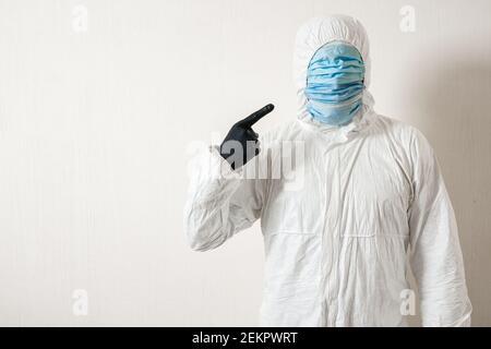 Ein Mann in einem Schutzanzug hing mit medizinischen Masken Posiert vor einer Wand Hintergrund zeigt verschiedene Gesten mit seinem Finger Stockfoto