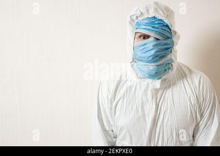 Ein verängstigter Mann in einem Schutzanzug, der mit medizinischen Masken aufgehängt ist, zeigt Schrecken an einer weißen Wand. Die Schrecken der Epidemie, die Gefahr des coro Stockfoto