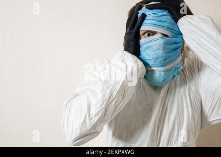 Ein verängstigter Mann in einem Schutzanzug, der mit medizinischen Masken aufgehängt ist, zeigt Schrecken an einer weißen Wand. Die Schrecken der Epidemie, die Gefahr des coro Stockfoto