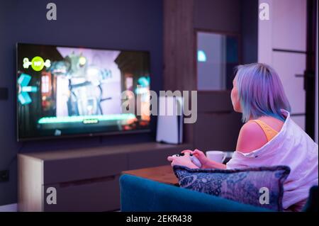 Schöne blauhaarige Mädchen hält Gaming-Contorller spielen Videospiele zu Hause. Rückansicht. Stockfoto