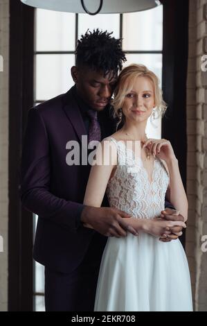 Interrassisches Paar, das vor einem großen Fenster steht. Fotoshooting mit Braut im Interracial-Sex. Kaukasische Frau, die mit afrikanischer dunkelhäutig heiratet Stockfoto