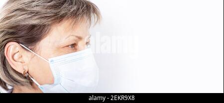 Ältere kaukasische Frau trägt medizinische Gesichtsmaske und schaut nach links. Bannerformat. Medizinischer weißer Hintergrund im Krankenhaus. Speicherplatz kopieren. Stockfoto