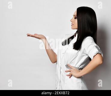 Junge hübsche Frau Arzt Krankenschwester im medizinischen Kleid steht seitlich Die Kamera hält die Hand hoch und schaut auf ihre offene Handfläche Stockfoto