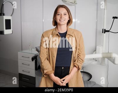 Junge Frau lächelt zur Kamera, während sie in der Zahnarztpraxis posiert. Charmante Frau zum Ausdruck positive Emotionen, während im Stomatologen Schrank stehen. Konzept der Zahnmedizin, Medizin und Gesundheitswesen. Stockfoto