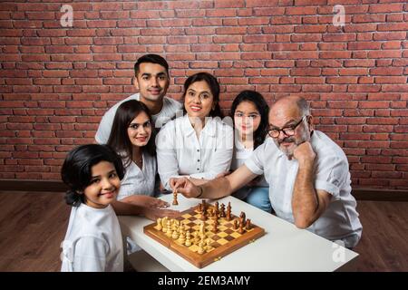 Happy Indian Asian Multigenerational Familie von sechs spielen Schach oder Shatranj, die ein beliebtes Brettspiel ist, trägt weiße Tücher gegen rote Ziegel Wal Stockfoto