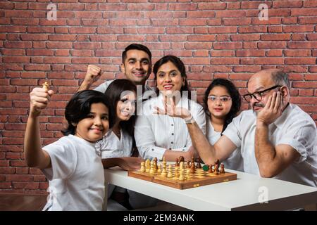 Happy Indian Asian Multigenerational Familie von sechs spielen Schach oder Shatranj, die ein beliebtes Brettspiel ist, trägt weiße Tücher gegen rote Ziegel Wal Stockfoto