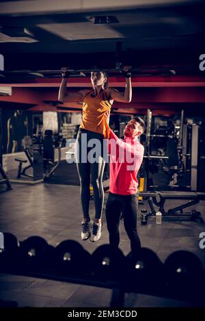 Fokussierte motivierte junge schlanke sportliche Fitness-Frau tun Pull-ups Übungen an einer Bar in der Turnhalle, während sie stark Muskulöses persönliches Stehen hinter einem Stockfoto