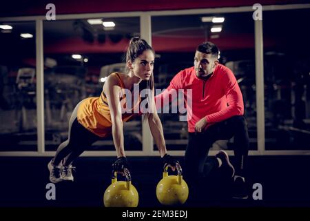 Nahaufnahme der motivierten fokussierten attraktiven jungen Frau tun Push Ups trainieren mit ihr auf den Kettlebells im Fitnessstudio Schöner muskulöser Personal Trainer Stockfoto