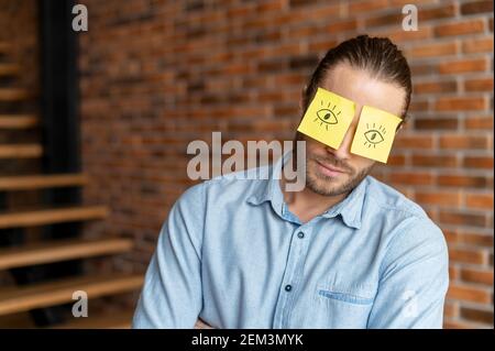 Müder Mann mit gelben Aufklebern mit bemalten Augen auf dem Gesicht, schlafend am Arbeitsplatz, fleissig, leicht zur Seite gelehnt, in blau gekleidet Stockfoto