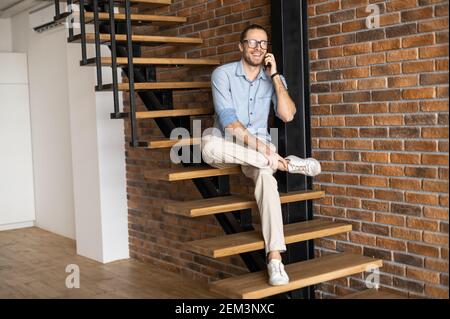 Schlanker Geschäftsmann Mann mit schwarzen Gläsern und Bart, einen Fuß zum anderen, sitzt auf einer Holztreppe, Loft-Stil innen, Durchführung der Arbeit Stockfoto