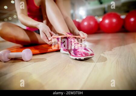 Nahaufnahme Fokus Ansicht von Sneakers und Hände während junge Form sportlich aktive Mädchen binden Schnürsenkel in der Turnhalle mit kleinen Hantel in der Nähe. Stockfoto