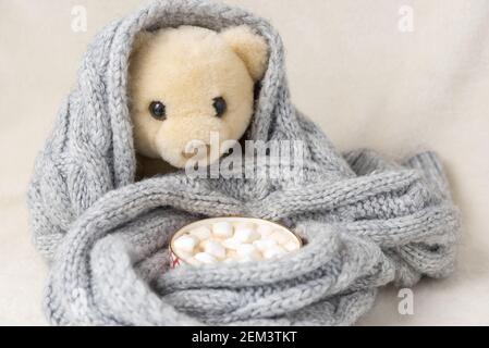 Ein weißer Teddybär ist in einen Schal gewickelt, neben einer Tasse heißer Schokolade mit Marshmallows in den Girlande-Lichtern Stockfoto