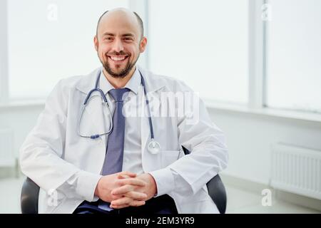 Lächelnder Arzt sitzt auf einem Stuhl und schaut auf die Kamera. Stockfoto