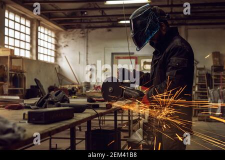 Seitenansicht des fokussierten gut aussehenden fleißigen Mannes mit Schutzmaske Arbeiten mit elektrischen Schleifwerkzeug auf der Stahlkonstruktion in Die Fabrik während Balken Stockfoto