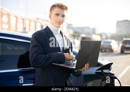 Nahaufnahme Porträt von jungen schönen blonden Mann in trendigen Outfit, arbeiten auf Laptop-pc in der Nähe Aufladen Elektroauto im Freien auf dem Hintergrund der verschwommen Stockfoto