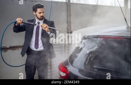 Nahaufnahme Porträt-Ansicht von stilvoll fokussierten bärtigen jungen fleißigen Selbstbewusster Mann im Anzug wäscht sein schwarzes Auto mit einem Wasserpistole auf der manuellen selbst-se Stockfoto