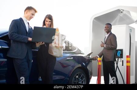 Lächelnder afrikanischer Mann in formeller Kleidung, stecken Stecker in Elektroauto zum Aufladen, Blick auf die Kamera und zeigt Daumen nach oben, während sein Geschäft Stockfoto