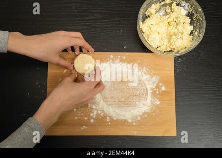 Frauenhände Formen Hüttenkäse-Pfannkuchen auf einem Holzbrett mit Mehl auf einem schwarzen Tisch. Stockfoto