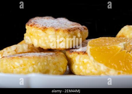 Süße Hüttenkäse-Pfannkuchen mit orangefarbenen Scheiben auf einem weißen Teller, Nahaufnahme an einer schwarzen Wand mit Platz für Kopien. Stockfoto