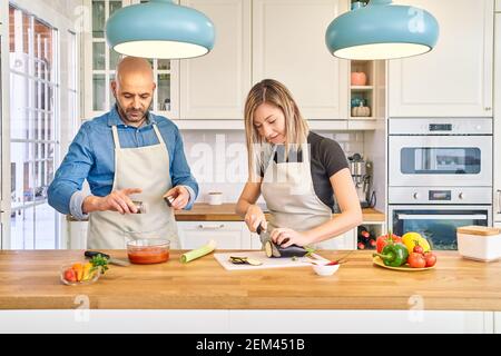 Ein glückliches junges Paar genießt und bereitet ein gesundes Essen in ihrer Küche zu. Sie schneidet Gemüse, er bereitet die Sauce. Kochen als Paar Stockfoto