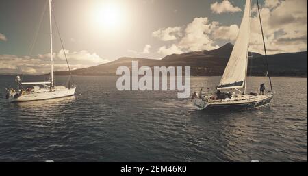 Sonne über Segelregatta auf Luxusyachten Luft. Filmische Seeslandschaft von Segelsport Sommer sonnigen Tag. Panorama Drohne aufgenommen. Majestätische Segelboote Rennen in Ocean Bay im Hafen von Brodick, Arran Island, Schottland Stockfoto