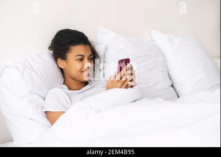 Glückliche junge Mixed-Race-Frau mit lockigen Haaren bedeckt mit Decke zu Hause im warmen Bett bleiben, im Chat mit Freunden auf dem Handy, flirten Stockfoto