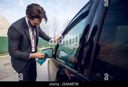 Nahaufnahme eines stylischen, konzentrierten und fleißigen jungen Geschäftsmannes, der den Rückspiegel seines schwarzen Autos mit einem blauen Mikrofasertuch putzt. Stockfoto