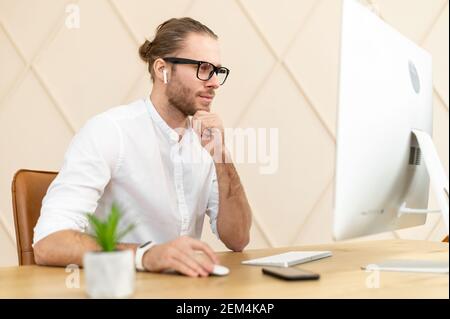 Reifer Mann, der am Computer sitzt, ernsthaft auf den Bildschirm schaut, im Internet sucht, weißes Hemd trägt, hellbraune Haare, Brille und Pferdeschwanz hat, Messaging mit Partnern, Denken Stockfoto