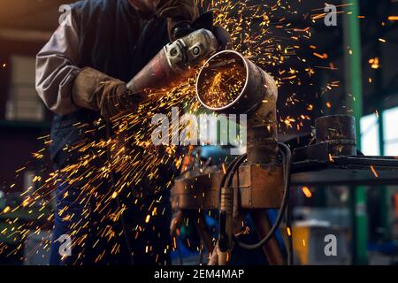 Side Nahaufnahme des professionellen fokussierten fleißigen Mann in Uniform Arbeiten an der Metallrohrskulptur mit einem elektrischen Grinder, während Funken fliegen Stockfoto