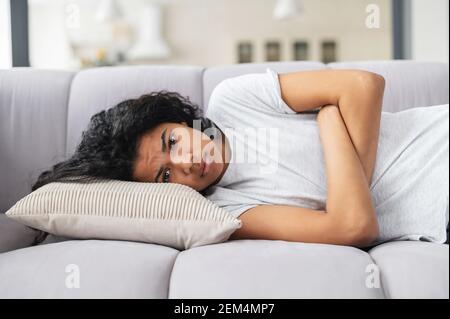 Depressive junge afroamerikanische Teenager-Mädchen mit lockigen Haaren auf der Couch mit gekreuzten Armen liegend, überwältigt, gestresst wegen unerfüllter Liebe oder brechen, wütend nach dem Kampf mit den Eltern Stockfoto