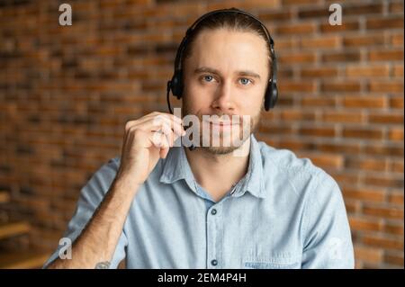 Selbstbewusster Bediener mit Headset, Blick auf die Kamera, in blauem Hemd gekleidet, bester Arbeiter, hat einen Charme, helfen Menschen ist seine Berufung, sprechen, hält seine Hand auf dem Mikrofon, verbessert den Klang Stockfoto