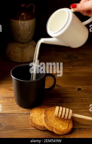 Eine weibliche Hand gießt Milch aus einem Krug in ein tasse mit Zwieback und Honig daneben Stockfoto